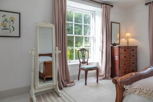 Un dormitorio con cama, una silla, un tocador, un espejo y una ventana con cortinas.