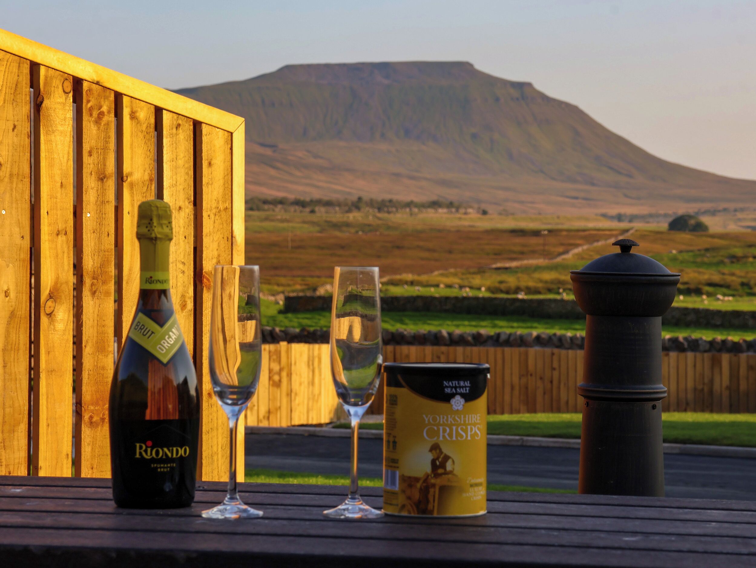 Una botella de vino Rionde Brut Organ, dos copas y una lata de Yorkshire Crisps sobre una mesa de madera con vistas a una colina y una valla de madera al fondo.