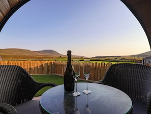 A table with a bottle and two glasses, overlooking a scenic landscape with hills and a wooden fence.