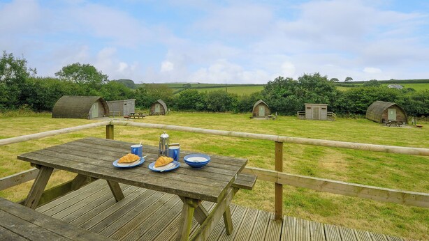 A wooden picnic table with plates of food and a metal kettle, set against a backdrop of rolling hills and several small wooden structures. - Hen House @ Penbugle Organic Farm (Liskeard)