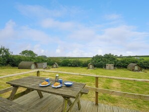A wooden picnic table with plates of food and a metal kettle, set against a backdrop of rolling hills and several small wooden structures. - Hen House @ Penbugle Organic Farm (Liskeard)
