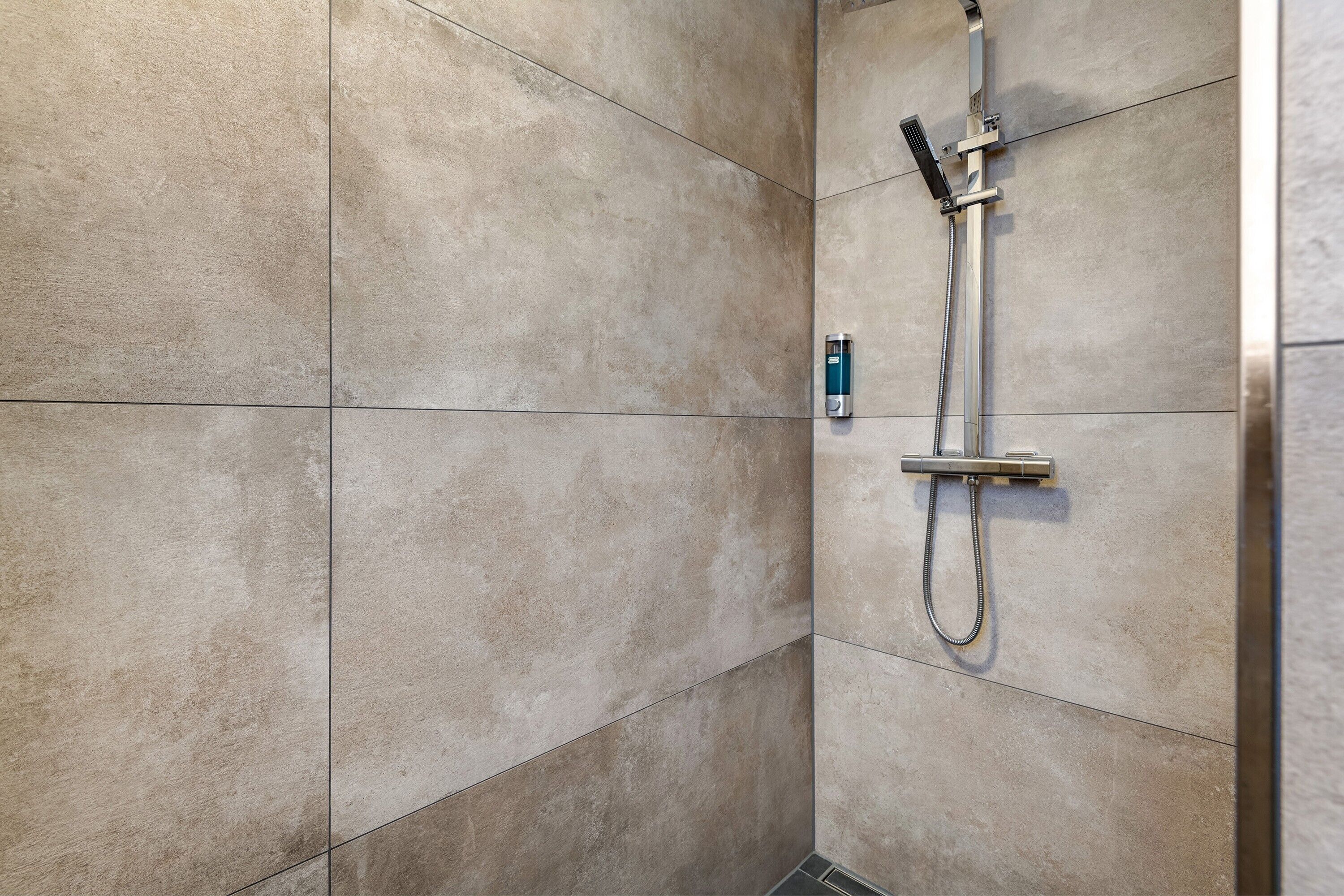 Une salle de bain avec des carreaux de couleur beige, une douchette à main et un robinet fixé au mur.
