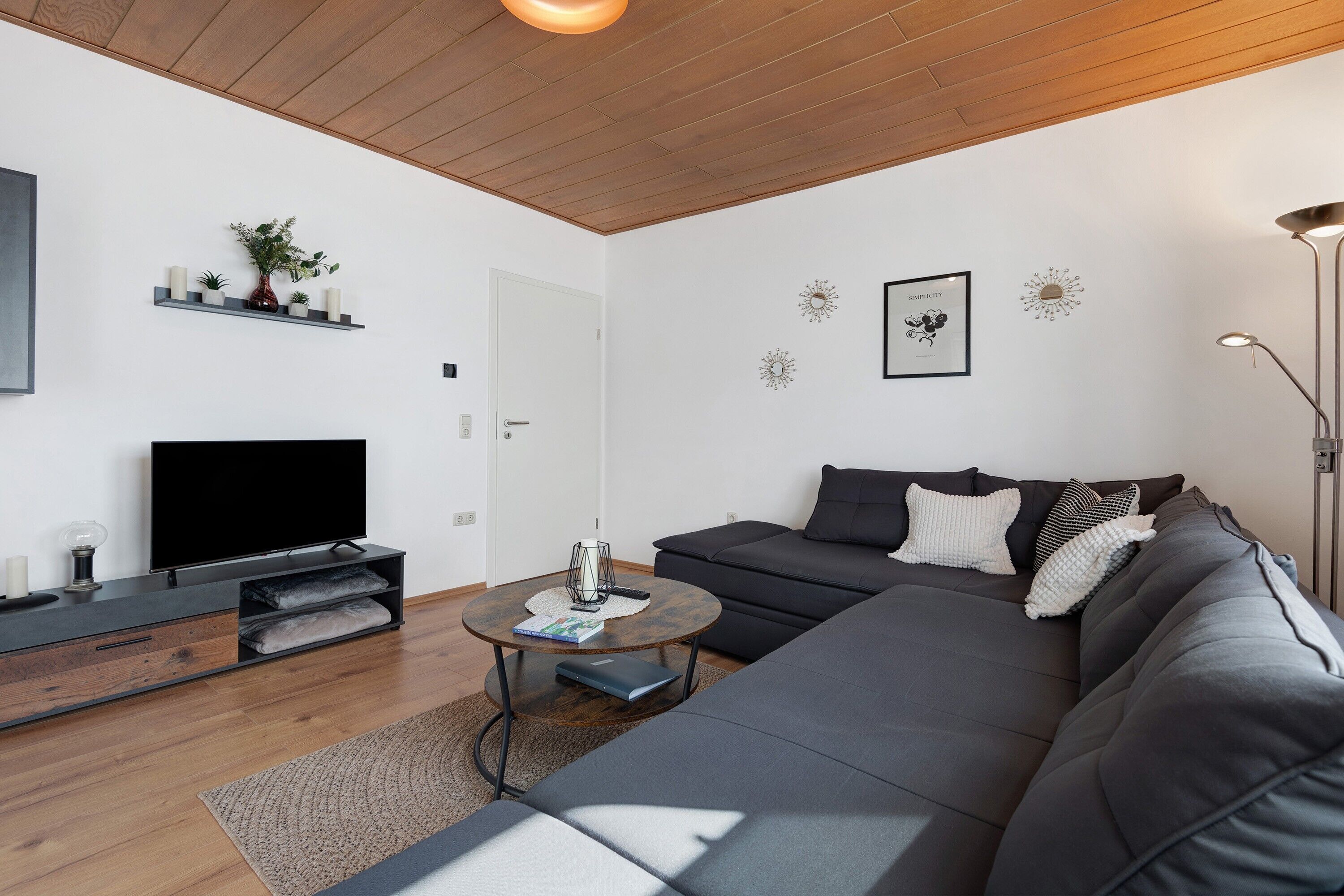 Ein modernes Wohnzimmer mit Flachbildfernseher, dunklem Sofa, Couchtisch und Holzboden.