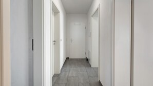 Un pasillo estrecho y bien iluminado, con paredes blancas y piso de baldosas grises, que conduce a una puerta al final.