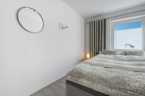 A bedroom with a bed, a bedside table with a lamp, a window with curtains, and a wall-mounted mirror.