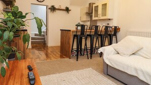 Een gezellige woonkamer met een bank, een salontafel en een tv-afstandsbediening. Een trap leidt naar een andere kamer, en er is een keukenhoek met stoelen en een aanrecht.