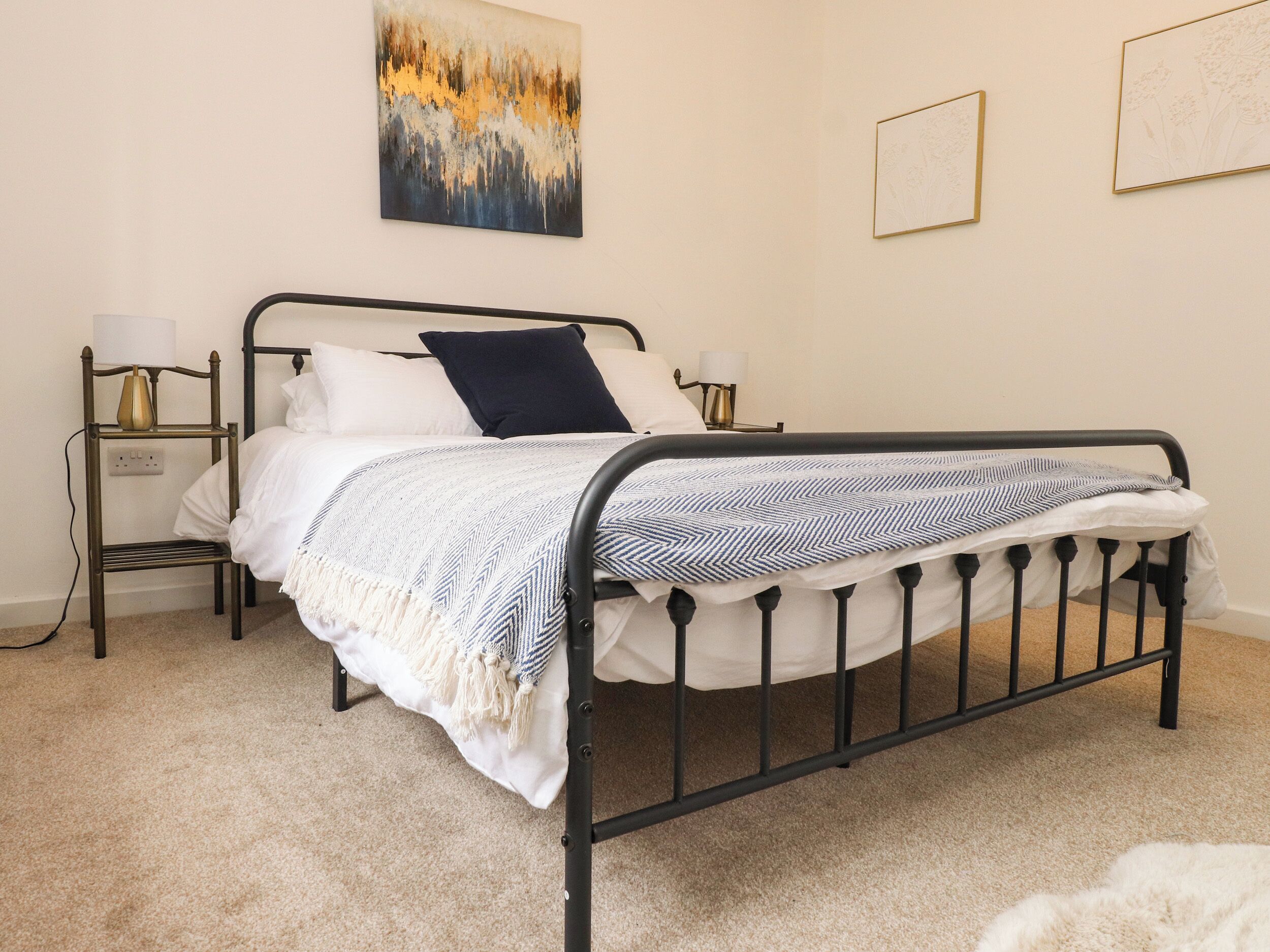 A neatly made bed with a white and blue striped blanket, a dark blue pillow, and a white throw. There are two bedside tables with lamps and a wall-mounted painting above the bed.