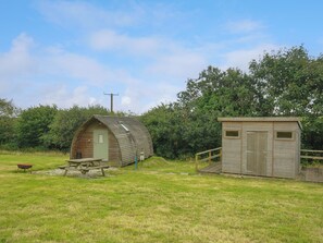A rustic cabin with a sloped roof and a small attached shed, surrounded by a grassy field and dense trees. - Fox Den @ Penbugle Organic Farm (Liskeard)