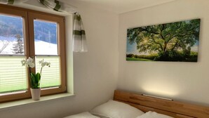 A bedroom with a wooden headboard, a window with blinds, a potted orchid, and a landscape painting. - Holiday Home 'Bei Schönwetter' with Mountain View, Private Terrace & Wi-Fi (Bleiberg)