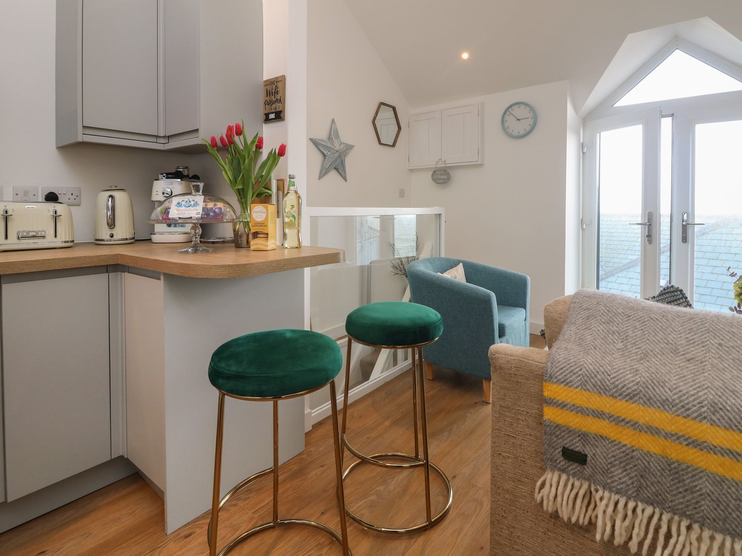 A modern kitchen with a wooden countertop, two green bar stools, a blue armchair, and a view of the sea through large windows.