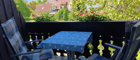 A balcony with two chairs and a table, surrounded by a black railing, with a view of buildings and trees.