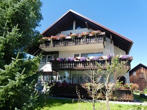 A two-story building with balconies adorned with flowers, a wooden fence, and a clear blue sky. - Apartment 'Diepold' with Mountain View, Shared Garden and Wi-Fi (Wasserburg (Bodensee))