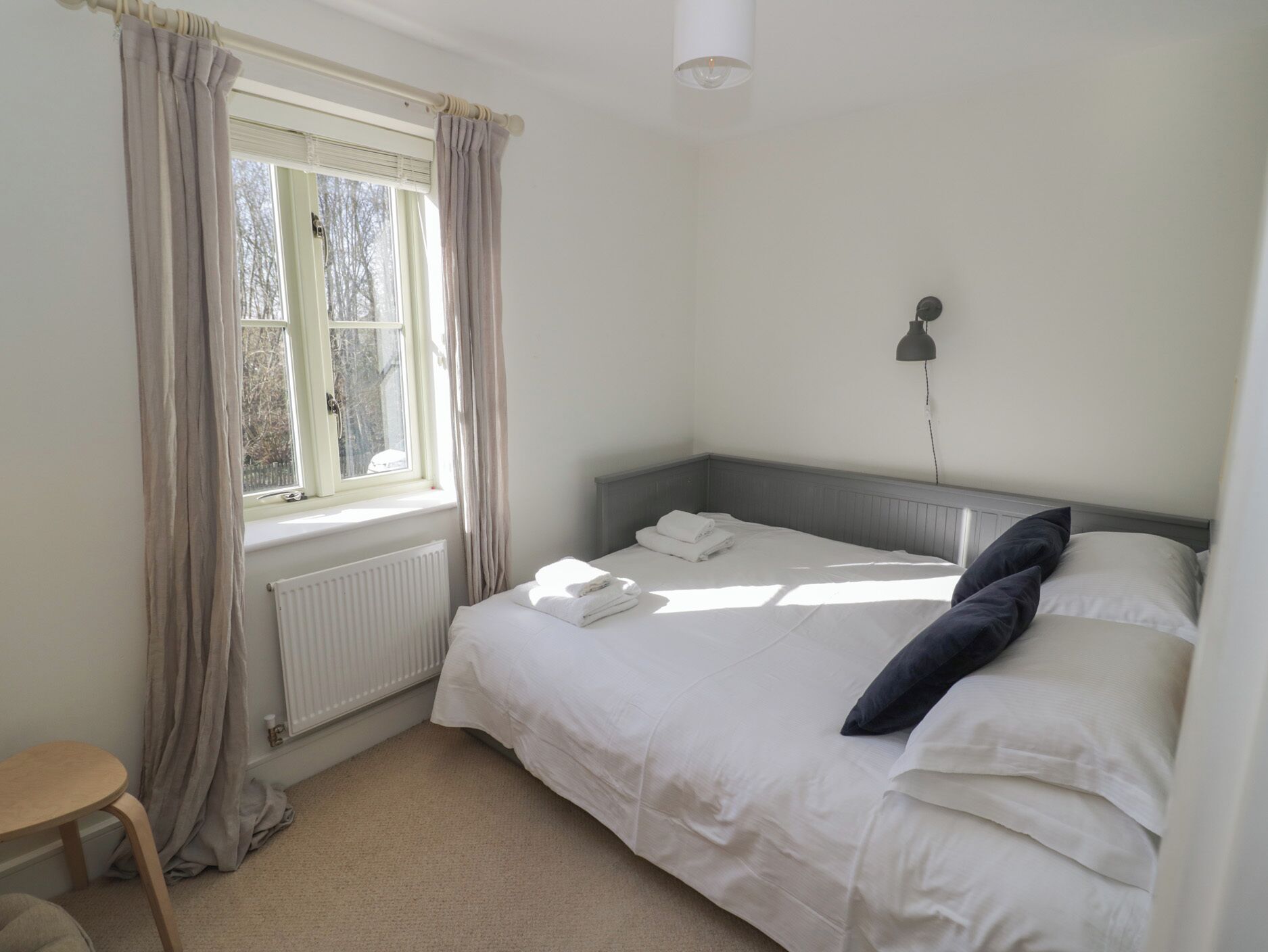 A bedroom with a bed, a window with curtains, a radiator, a bedside lamp, and a small table.