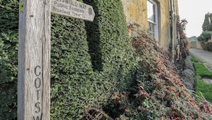 A wooden signpost with 'COTSWOLD WAY' and 'Broadway Tower' written on it, set against a backdrop of a building and lush greenery. - 2 Summers Way (Moreton-in-Marsh)