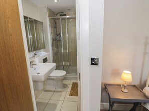 A modern bathroom with a glass shower enclosure, a white sink, and a toilet. There is a small table with a lamp next to the toilet. - 2 Summers Way (Moreton-in-Marsh)