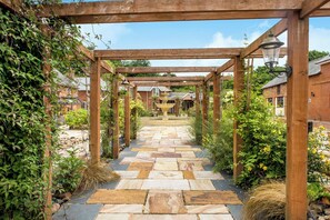 A stone pathway under a wooden pergola with a fountain in the center, surrounded by greenery and brick buildings. - Lime Cottage (Exeter)
