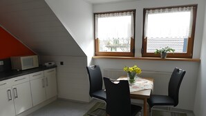 A compact kitchen with white cabinets, a microwave, and a dining table with chairs. There are two windows with curtains, a radiator, and a potted plant on the windowsill. - Apartment 'Diepold' with Mountain View, Shared Garden and Wi-Fi (Wasserburg (Bodensee))