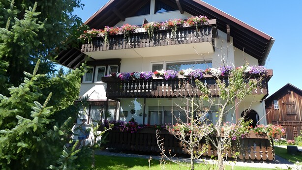 A two-story building with balconies adorned with flowers, a wooden fence, and a clear blue sky. - Apartment 'Diepold' with Mountain View, Shared Garden and Wi-Fi (Wasserburg (Bodensee))