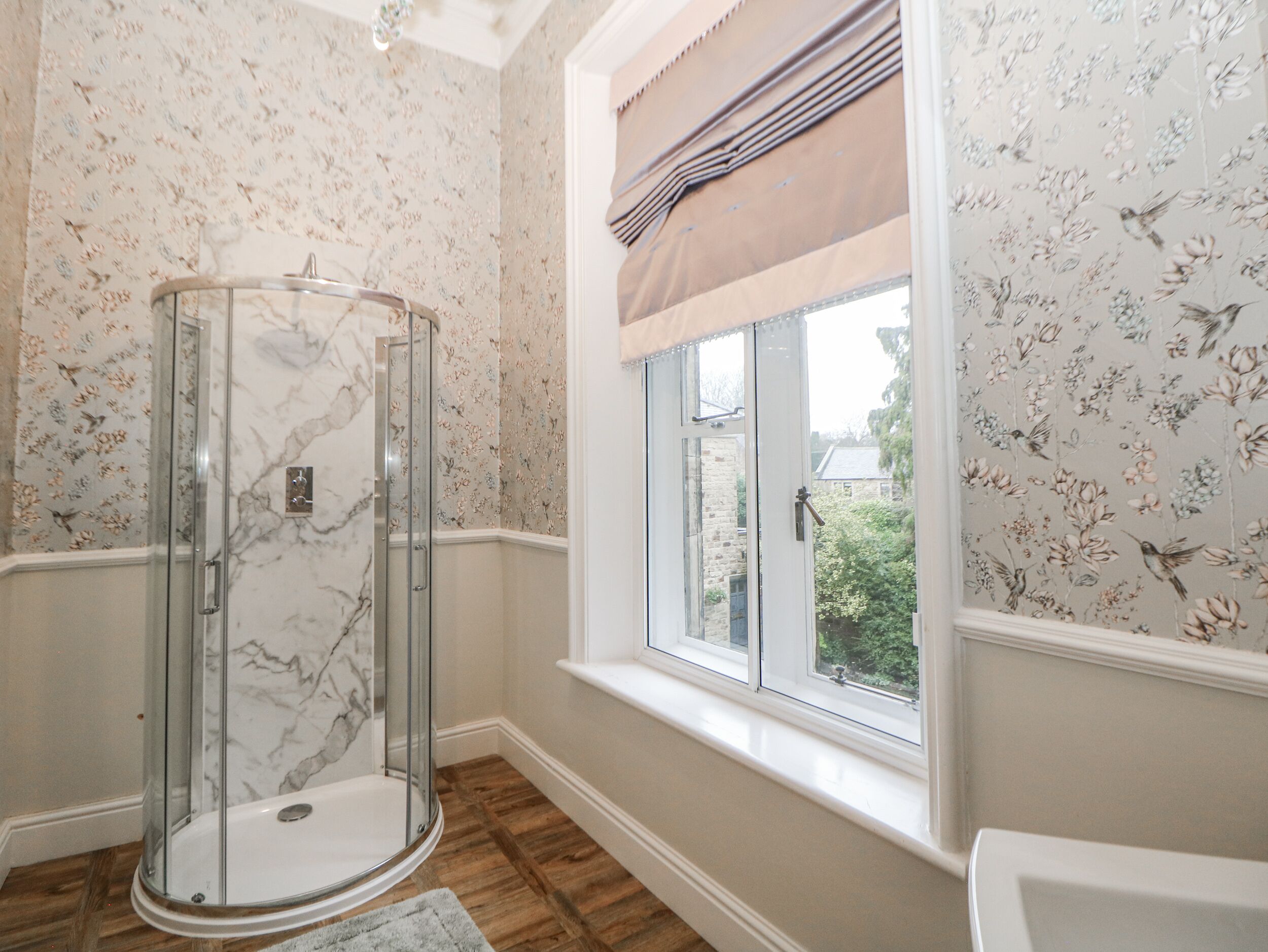 A bathroom with a glass shower enclosure, floral wallpaper, a window with a view of greenery, and a wooden floor.