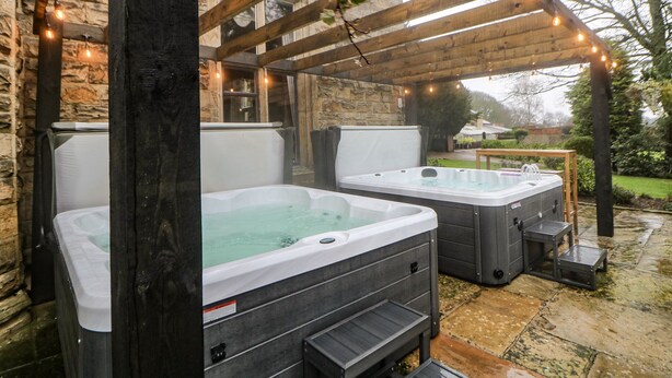 Dos jacuzzis al aire libre bajo una pérgola de madera con luces colgantes, frente a una pared de piedra y un jardín.