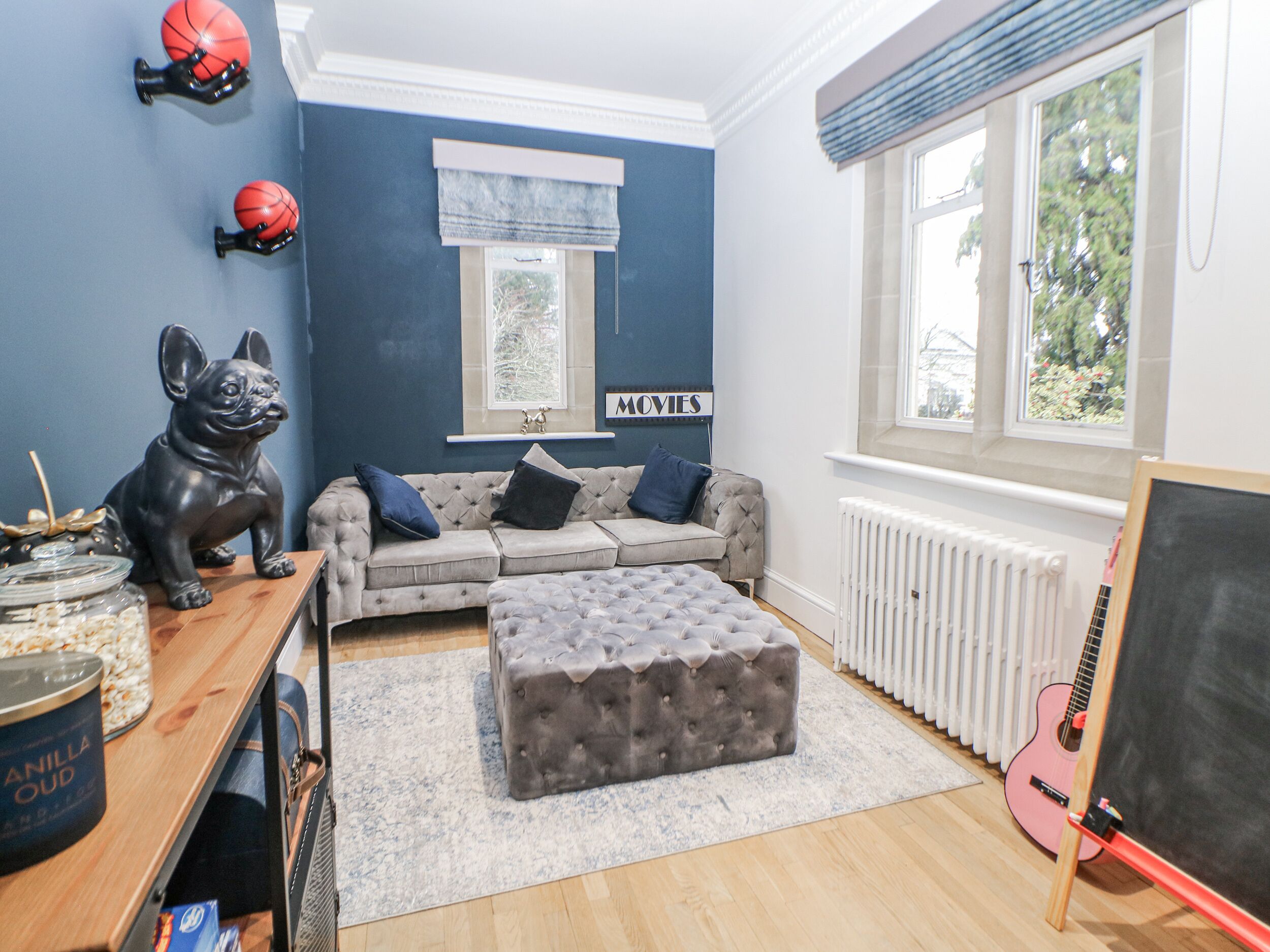A living room with a grey sofa, a tufted ottoman, a black dog figurine, a guitar, and a chalkboard.