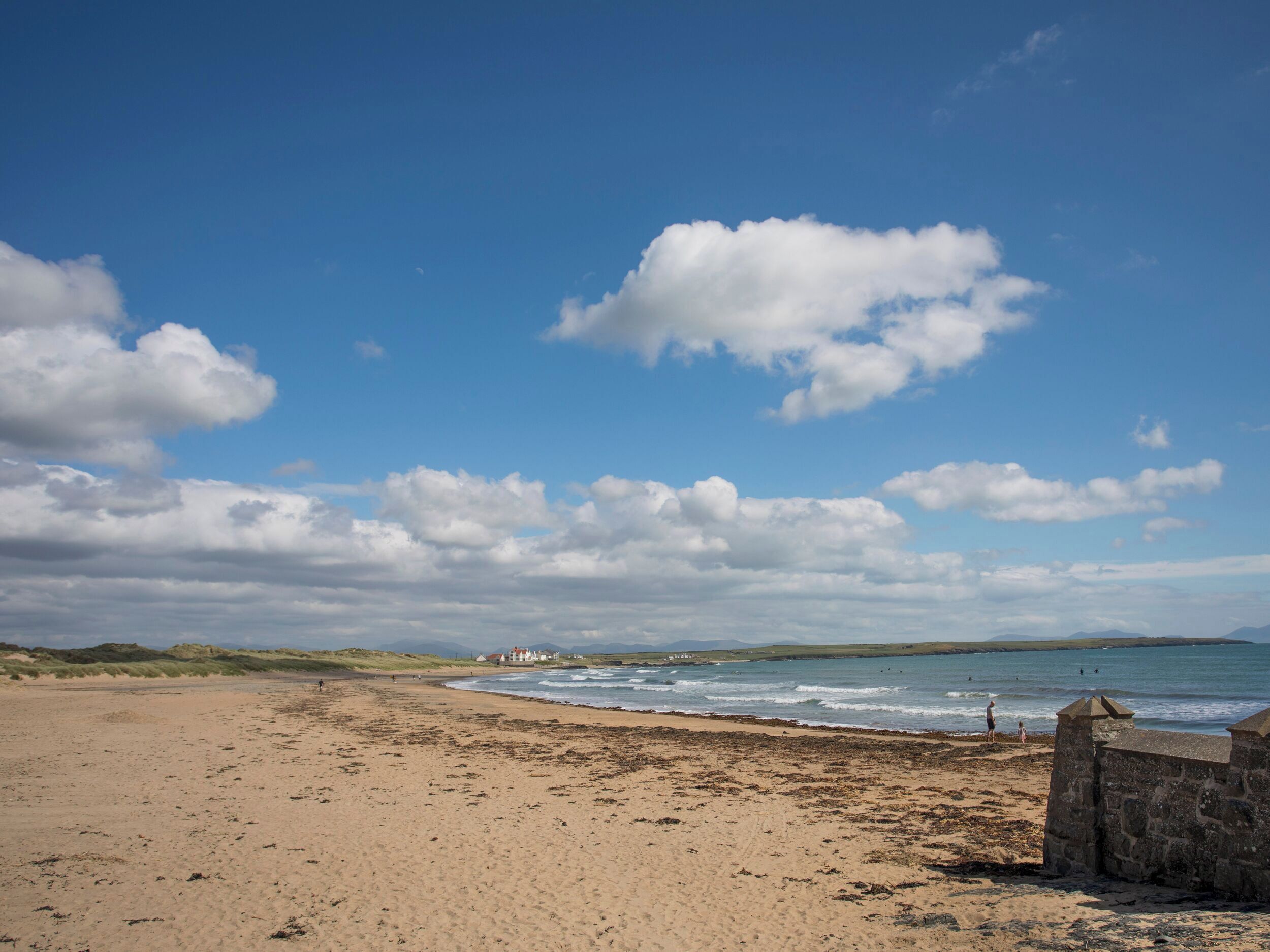 A beach with a stone wall, a few people, and a building in the distance.