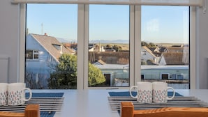 Une pièce dotée d’une grande fenêtre donnant sur un quartier résidentiel, une table avec deux tasses et une vue sur des maisons et des montagnes.