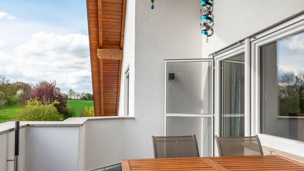 Un balcon avec une table et des chaises en bois, une porte-fenêtre coulissante et une vue sur la verdure.