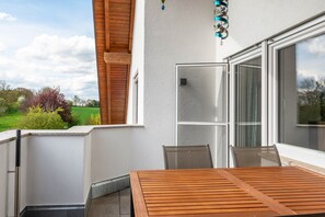 Een balkon met een houten tafel en stoelen, een schuifpui en uitzicht op groen.