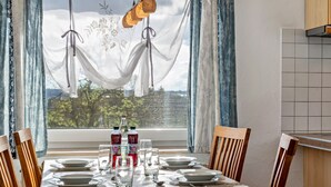 Une table à manger dressée pour quatre personnes, avec des assiettes, des verres et des bouteilles, une fenêtre avec des rideaux et une vue sur les arbres.