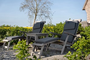 Deux fauteuils de jardin en bois avec des coussins, une petite table et un espace dallé de pierres agrémenté de verdure.