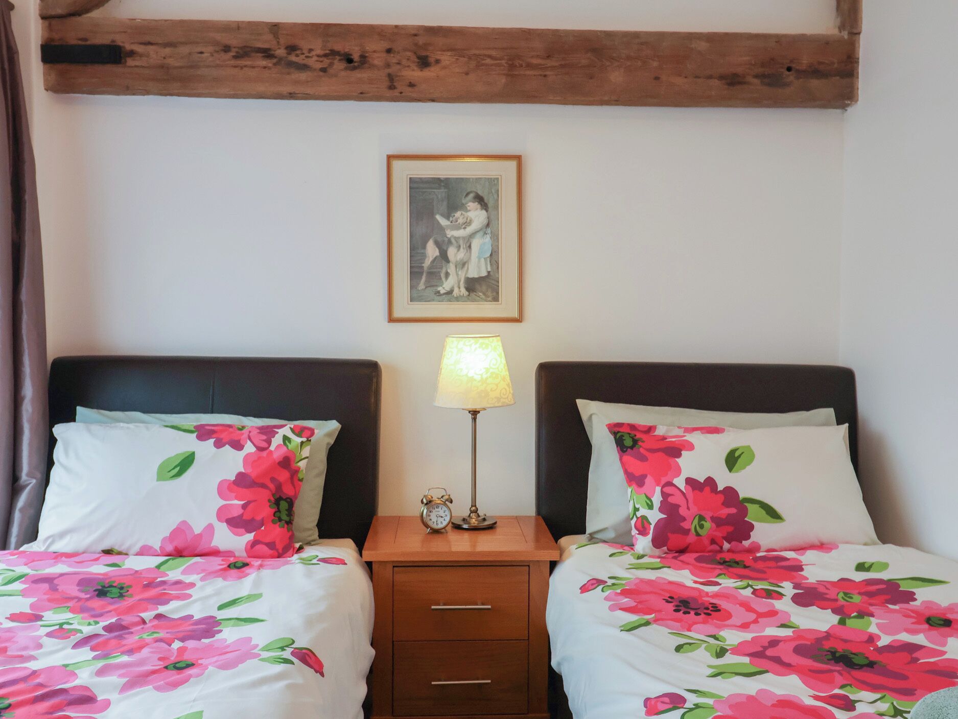 Een tweepersoonsbed met bloemenbeddengoed, een nachtkastje met een lamp, en een lijst met een afbeelding aan de muur.