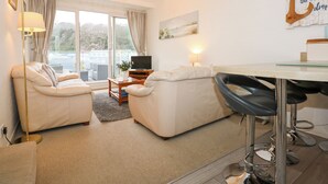 A modern living room with a sofa, armchairs, a coffee table, and a TV. There is a large window with curtains, a chandelier, and a wall-mounted artwork. - Anchor Watch (Porthmadog)