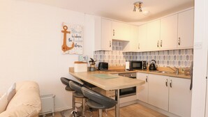 A compact kitchen with a wooden countertop, a microwave, a kettle, and a toaster. There are two bar stools and a small table. - Anchor Watch (Porthmadog)