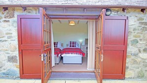 Un bâtiment en pierre avec une porte en bois donnant sur une chambre à coucher équipée de deux lits, d’une table de chevet et d’une lampe.