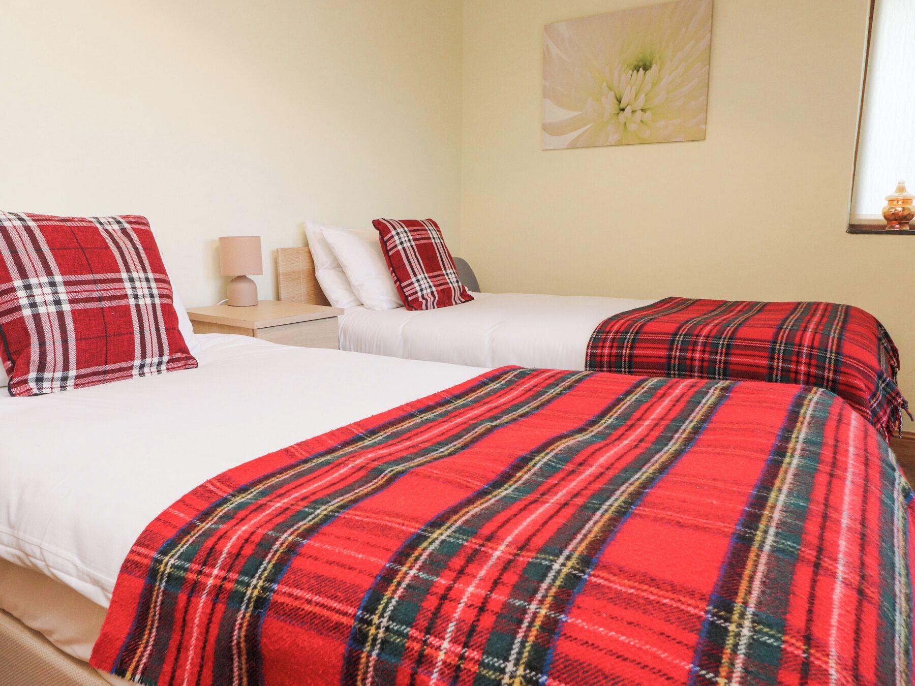 Habitación de hotel con dos camas, cada una con una manta a cuadros rojos y ropa de cama blanca. Se ve una mesita de noche con una lámpara y una obra de arte colgada en la pared.