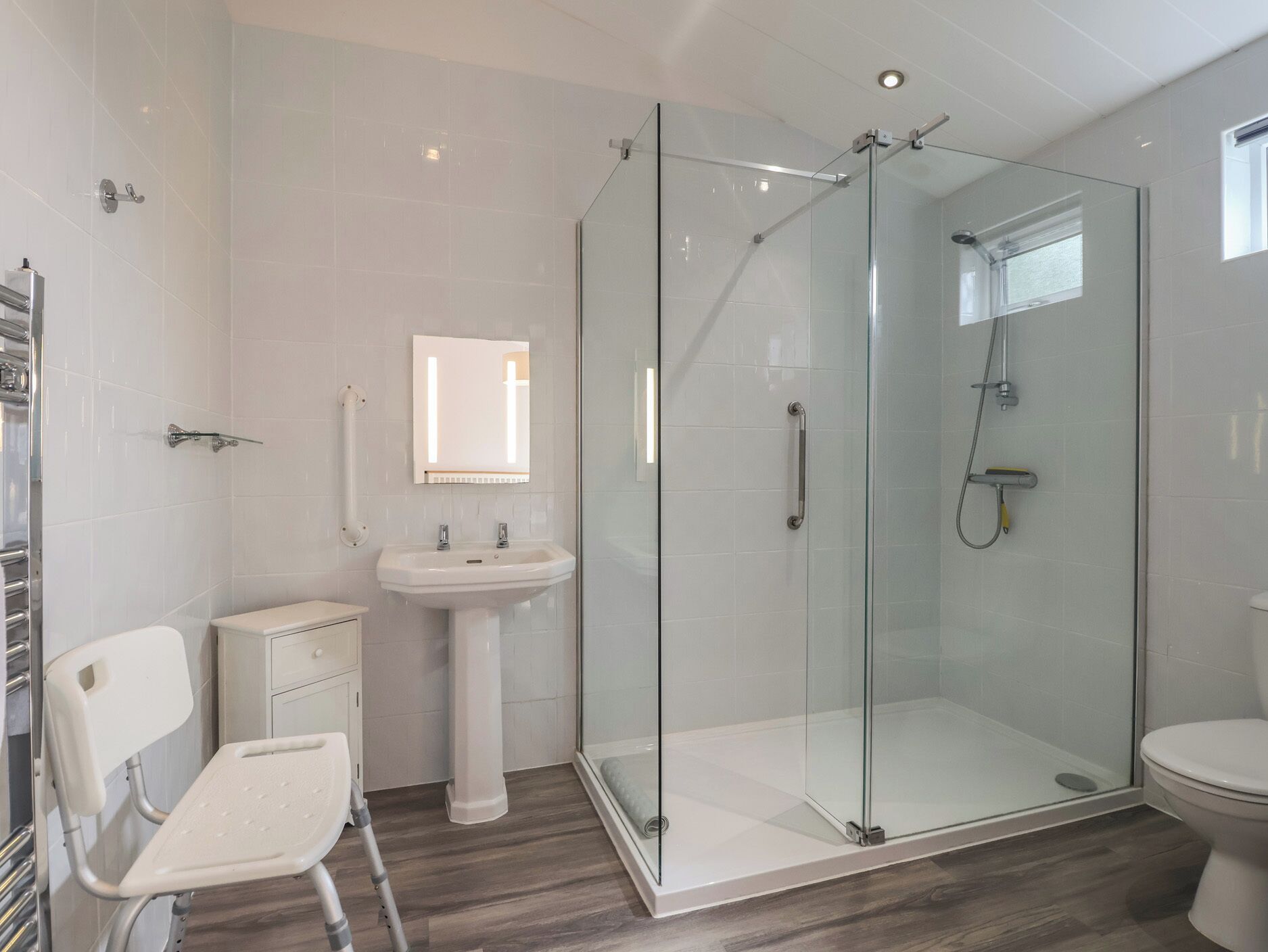 A modern bathroom with a glass-enclosed shower, a pedestal sink, a white toilet, and a white chair.