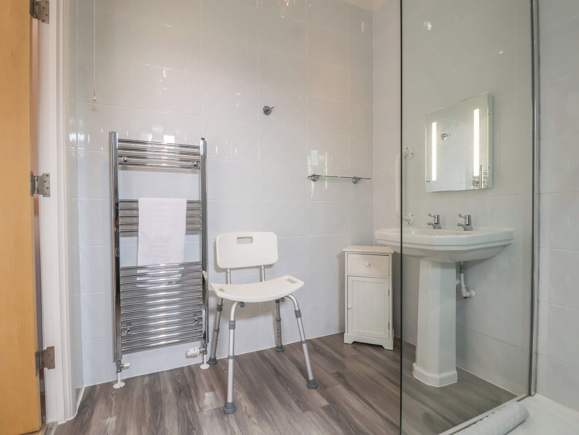 Een badkamer met een douchecabine van glas, een witte wastafel en een witte stoel.