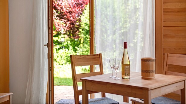 A wooden dining table with a bottle and glasses, a chair, and a view of greenery through a window. - Apartment 'Marie' with Private Terrace and Wi-Fi (Bodman-Ludwigshafen)