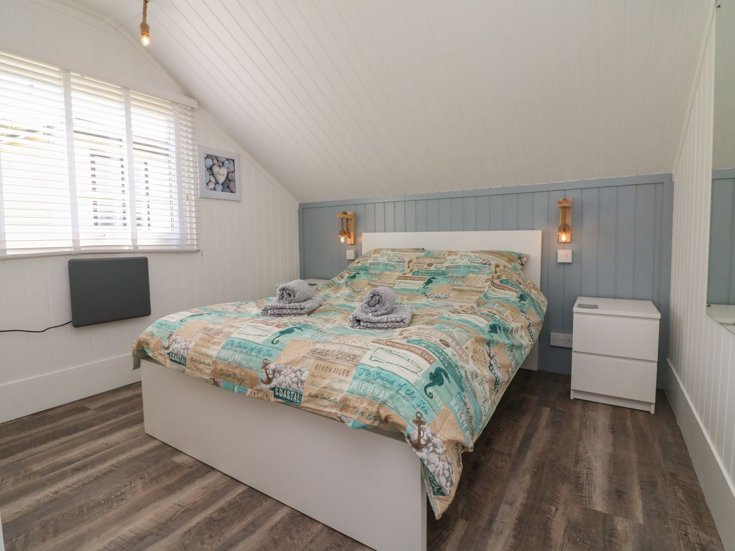 Un dormitorio con techo inclinado, una cama con cubrecama de temática costera, lámparas de noche, una mesita de noche pequeña y un radiador.