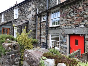 Une petite maison en pierre avec une porte rouge, des fenêtres blanches et un chemin de pierre menant à l’entrée.