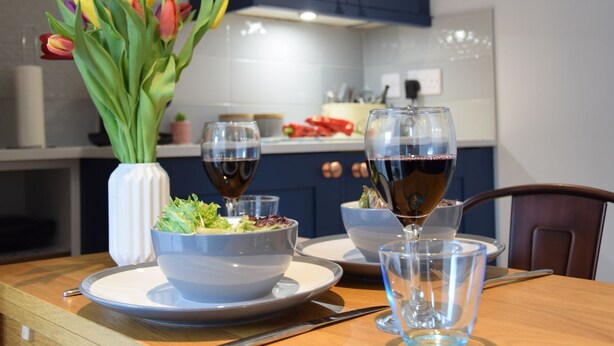 A modern kitchen with dark blue cabinets, a white countertop, and a dining table set with a salad bowl, plates, and wine glasses.