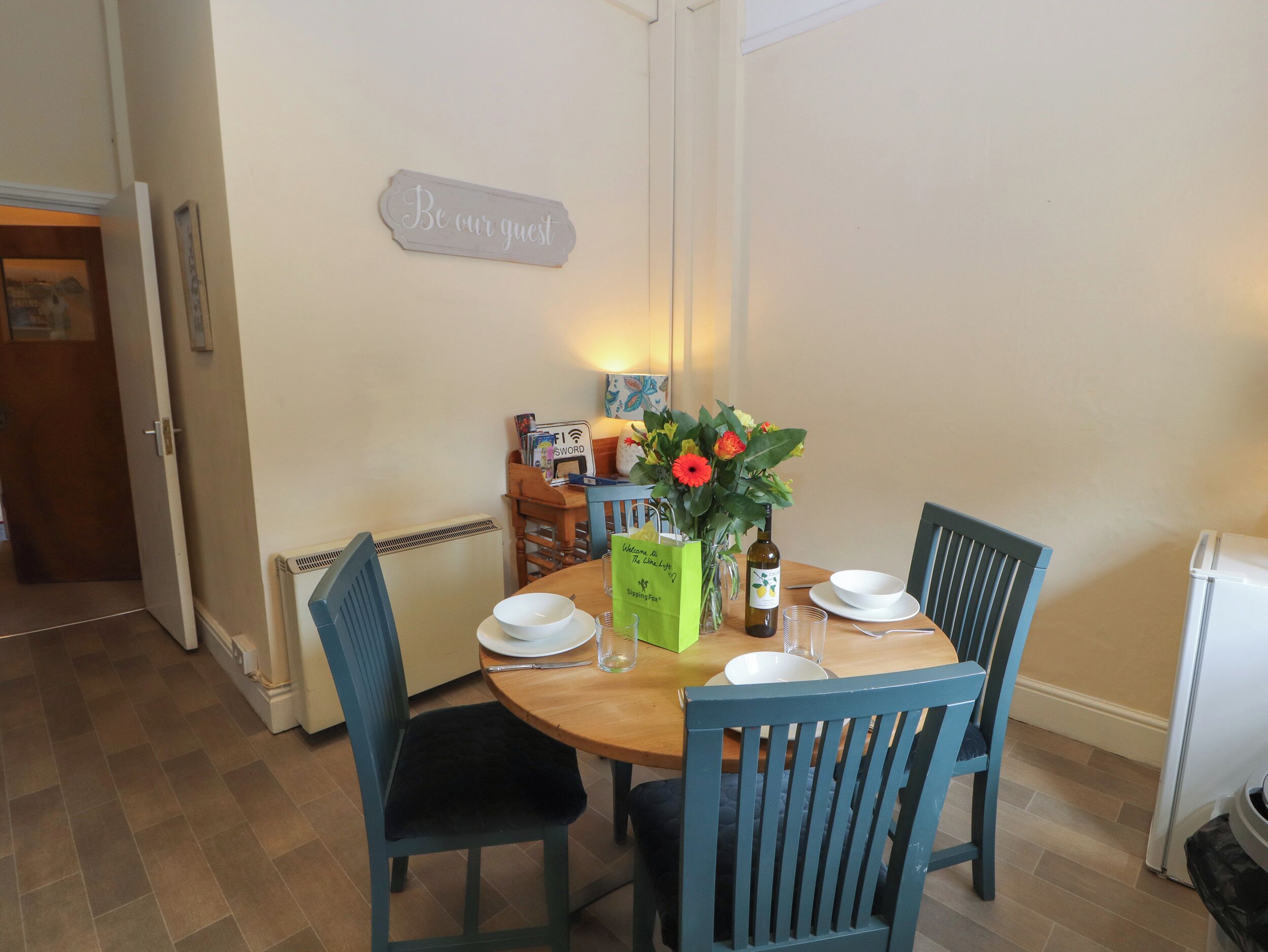 A dining area with a wooden table, four chairs, a vase with flowers, a bottle, and a green bag.