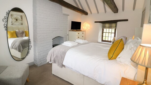 A bedroom with a white brick wall, a bed with white bedding and a yellow pillow, a wooden nightstand with a lamp, a dresser, and a television on a wooden stand. - Edmunds Cottage (Bedale)