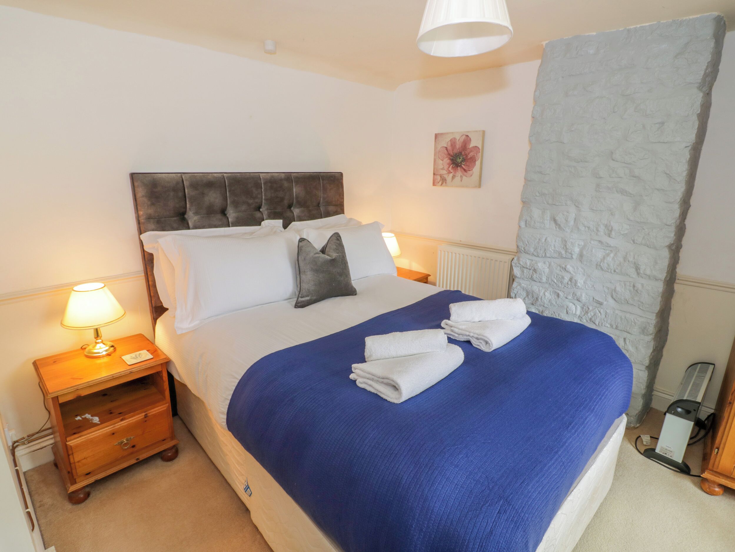 A neatly made bed with a blue blanket, a wooden nightstand with a lamp, and a radiator.