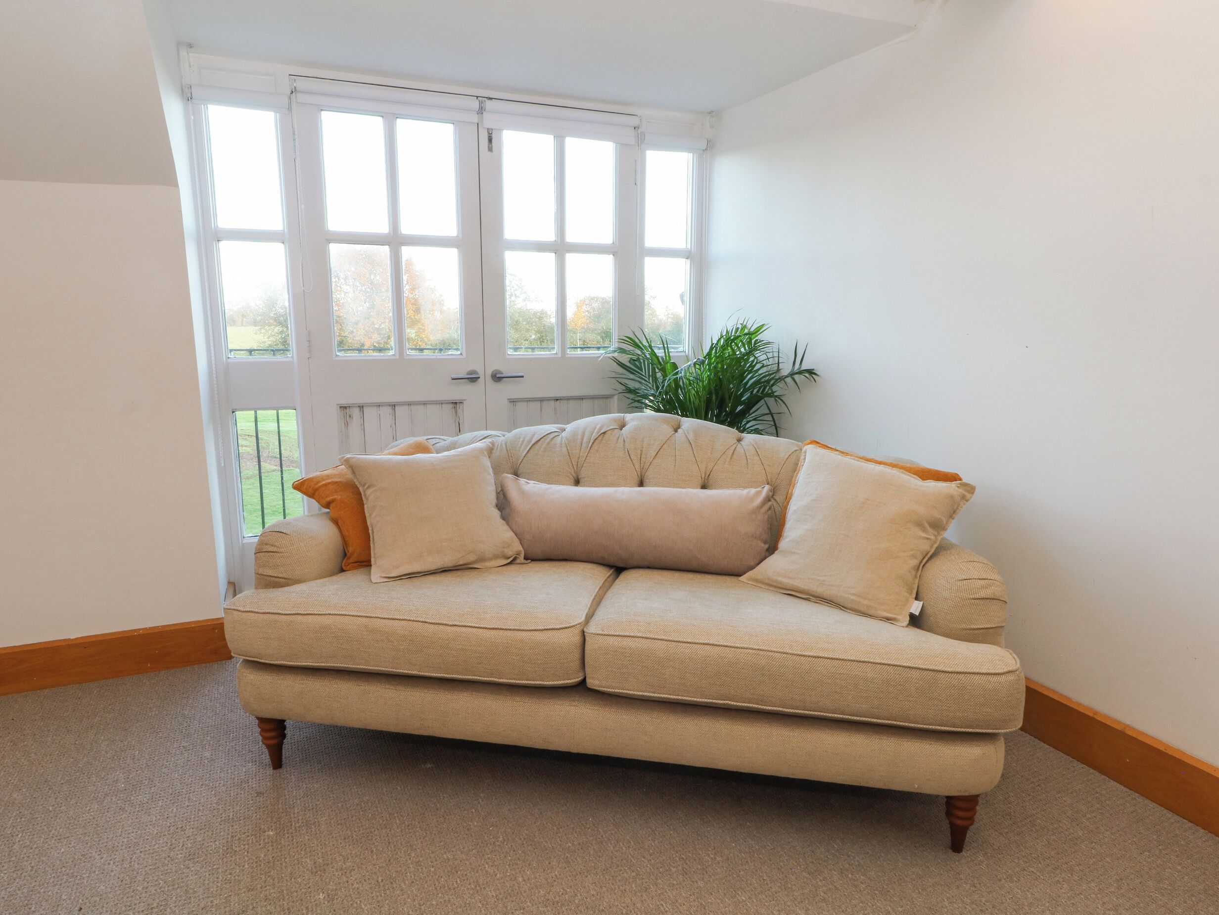 A beige sectional sofa with wooden legs in a corner of a room with white walls and a large window.
