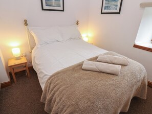 A neatly made bed with white linens, a fluffy beige throw blanket, and two folded towels. There is a bedside lamp on a wooden nightstand beside the bed.