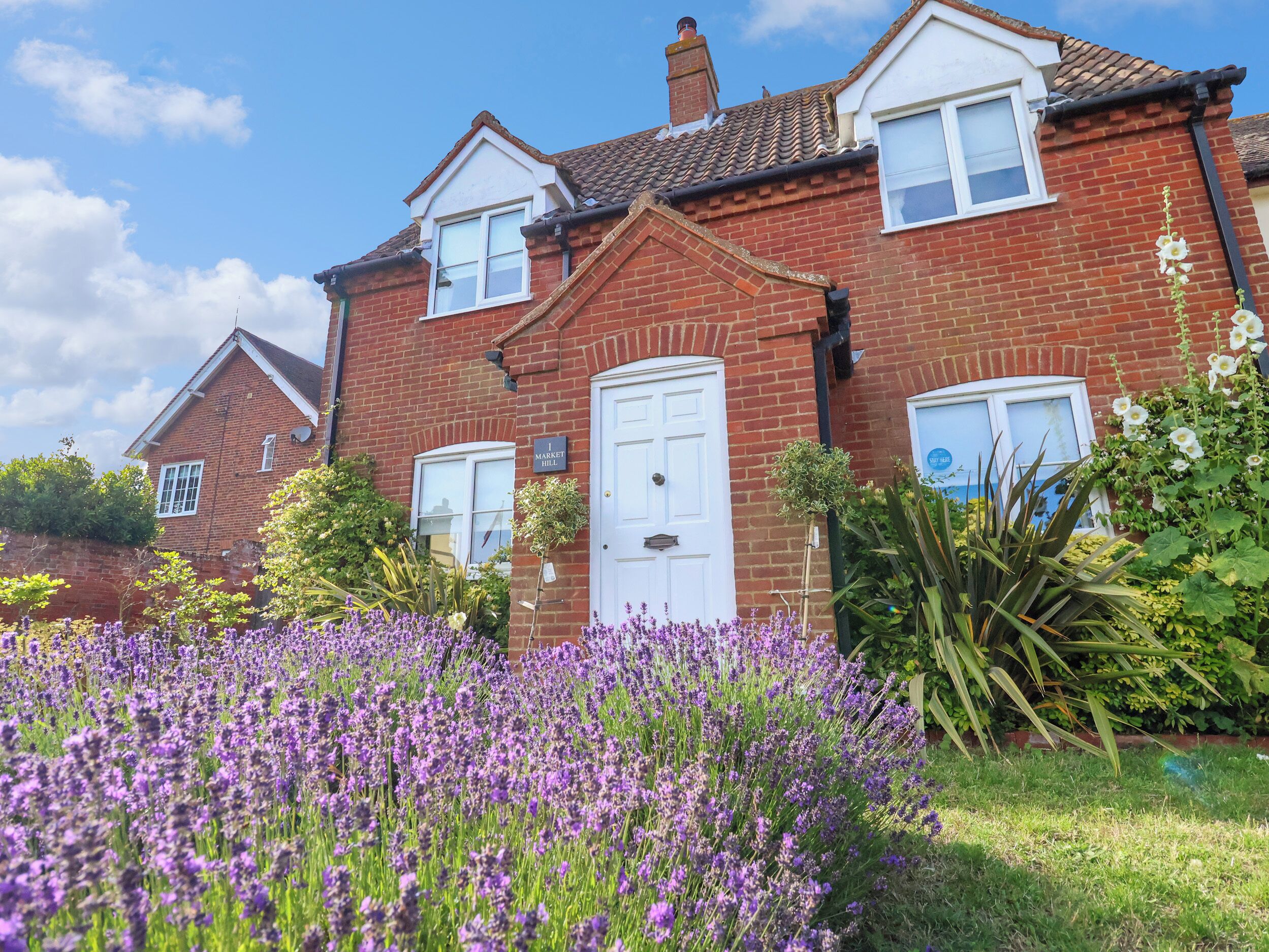 A two-story brick house with a white front door, two windows, and a small garden with purple flowers.