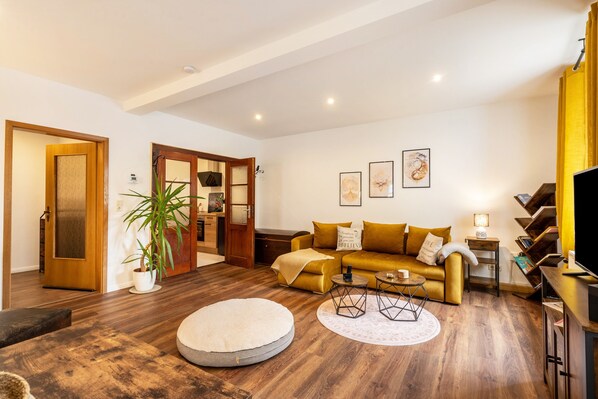 Una sala de estar con sofá, mesa de centro, reposapiés redondo, estantería y televisor.