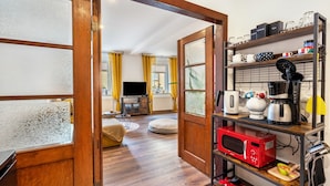 Una cocina moderna con microondas, cafetera y juego de té en estantes, un televisor al fondo y una puerta de madera que da a otra habitación.